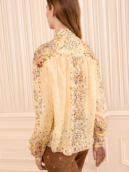 A woman shown from the back wearing a long-sleeve cream blouse with a delicate multicolor floral print and gathered shoulder details, paired with brown pants, set against a cream paneled wall.
