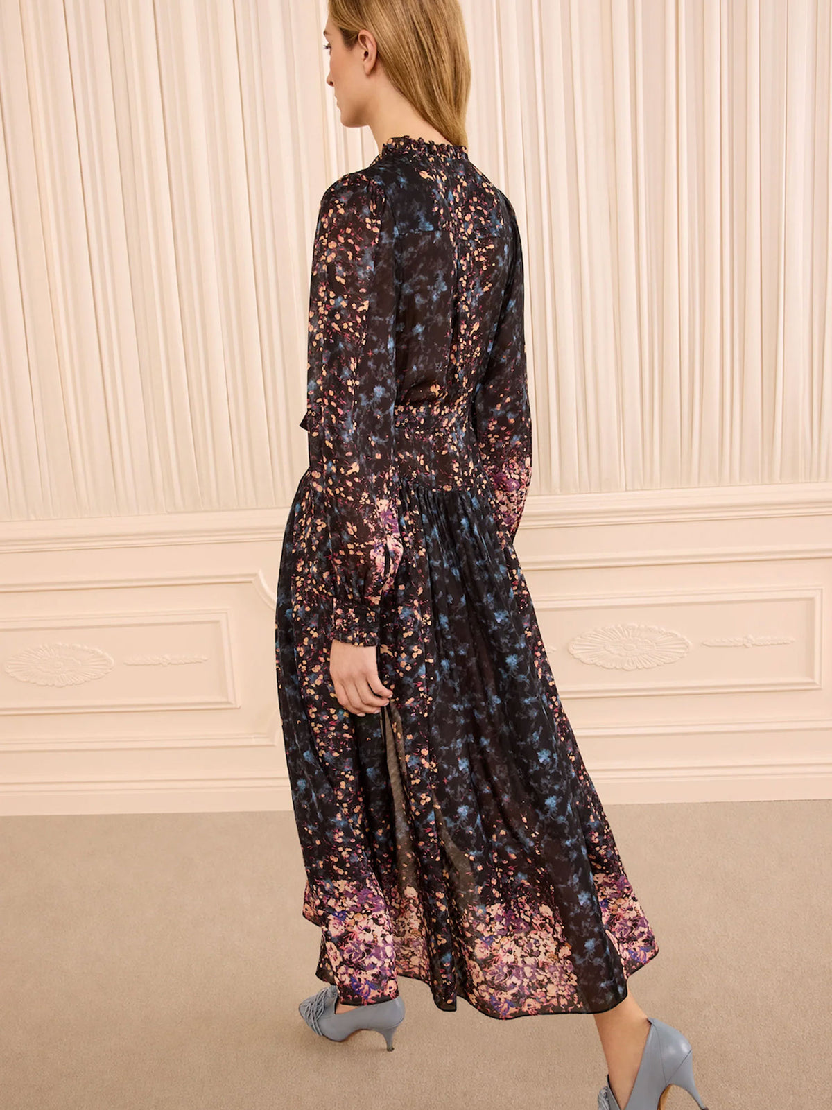A woman wearing a long-sleeved midi dress with a dark base and a dense floral pattern in purple, pink, and blue, photographed from behind in an elegant room with cream paneled walls.