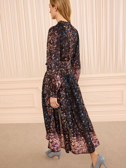 A woman wearing a long-sleeved midi dress with a dark base and a dense floral pattern in purple, pink, and blue, photographed from behind in an elegant room with cream paneled walls.