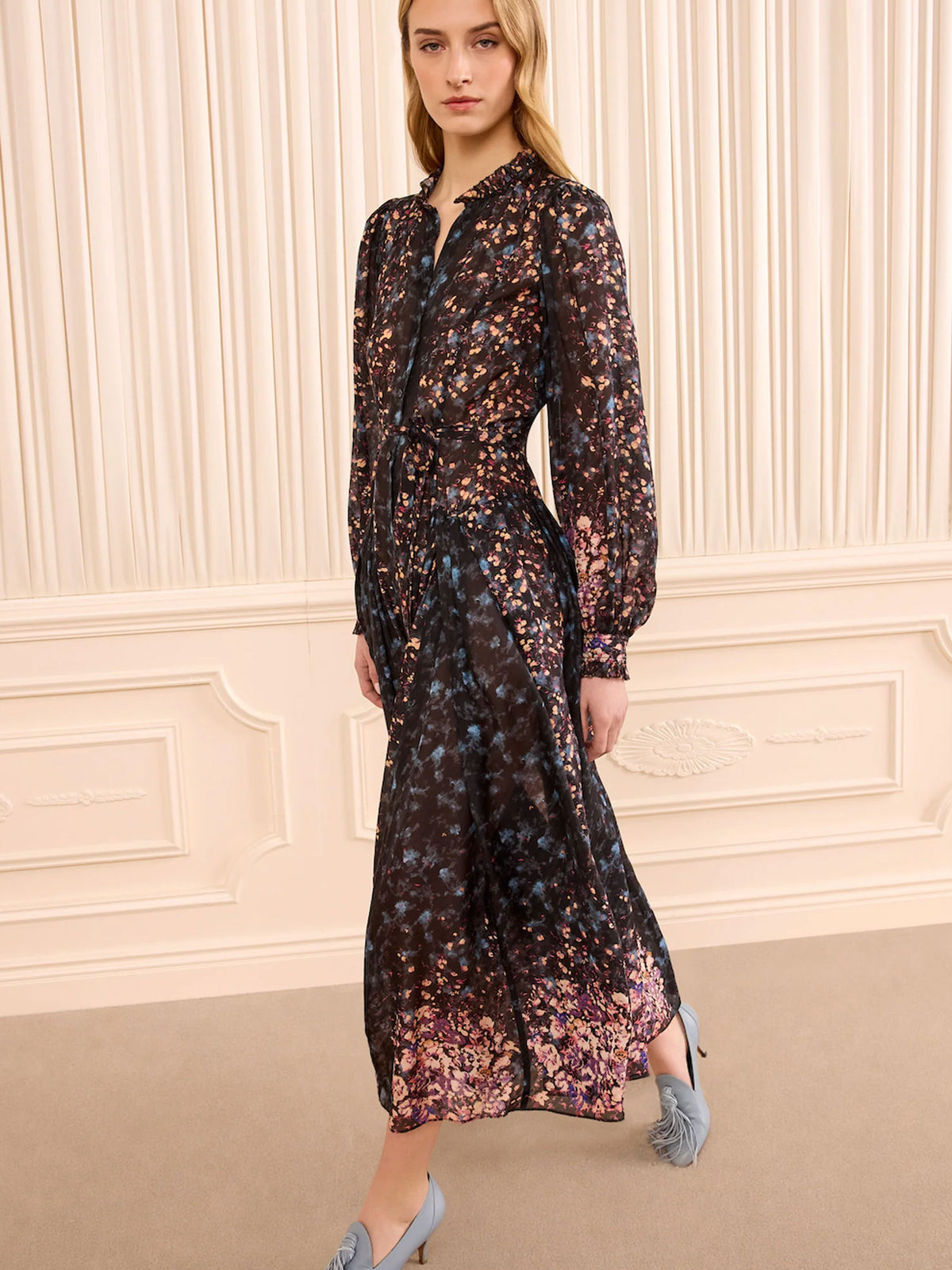 A woman models a long-sleeved midi dress with a dark base and a dense, multicolored floral print in shades of pink, purple, blue, and gold. The dress features a high collar, button-up front, and a matching self-tie belt at the waist.