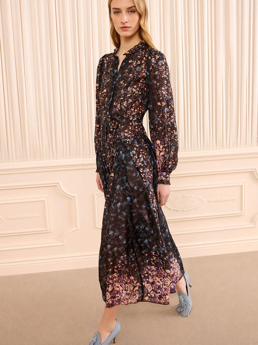 A woman models a long-sleeved midi dress with a dark base and a dense, multicolored floral print in shades of pink, purple, blue, and gold. The dress features a high collar, button-up front, and a matching self-tie belt at the waist.