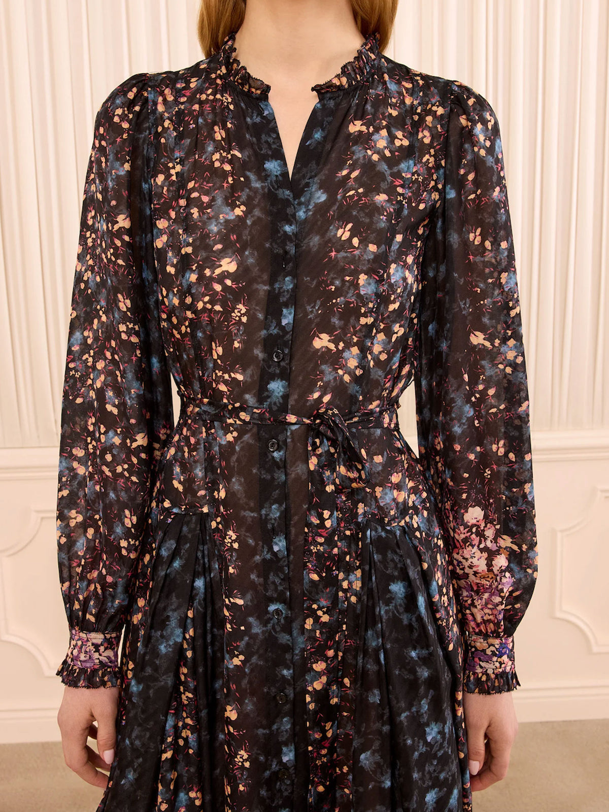 A woman wearing a long-sleeve black button-front dress with a multicolor floral print in blue, pink, and orange, featuring a tie waist and ruffled collar, set against a cream paneled wall.