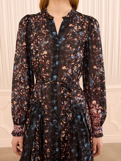 A woman wearing a long-sleeve black button-front dress with a multicolor floral print in blue, pink, and orange, featuring a tie waist and ruffled collar, set against a cream paneled wall.