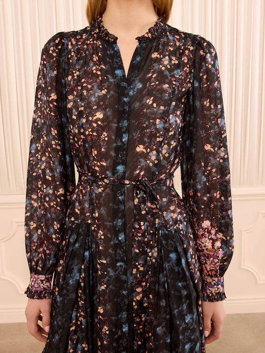A woman wearing a long-sleeve black button-front dress with a multicolor floral print in blue, pink, and orange, featuring a tie waist and ruffled collar, set against a cream paneled wall.