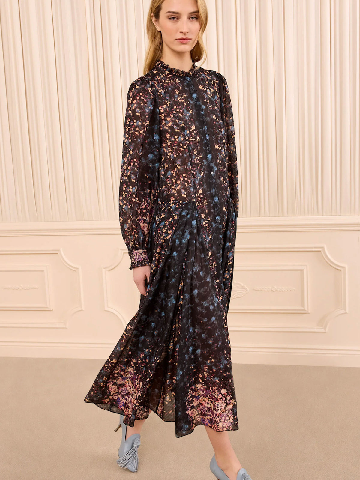 A woman models a long-sleeved, high-neck midi dress with a dark base and a dense floral print in pink, purple, and blue. She is wearing light blue pointed-toe flats, standing in front of a cream-colored paneled wall.