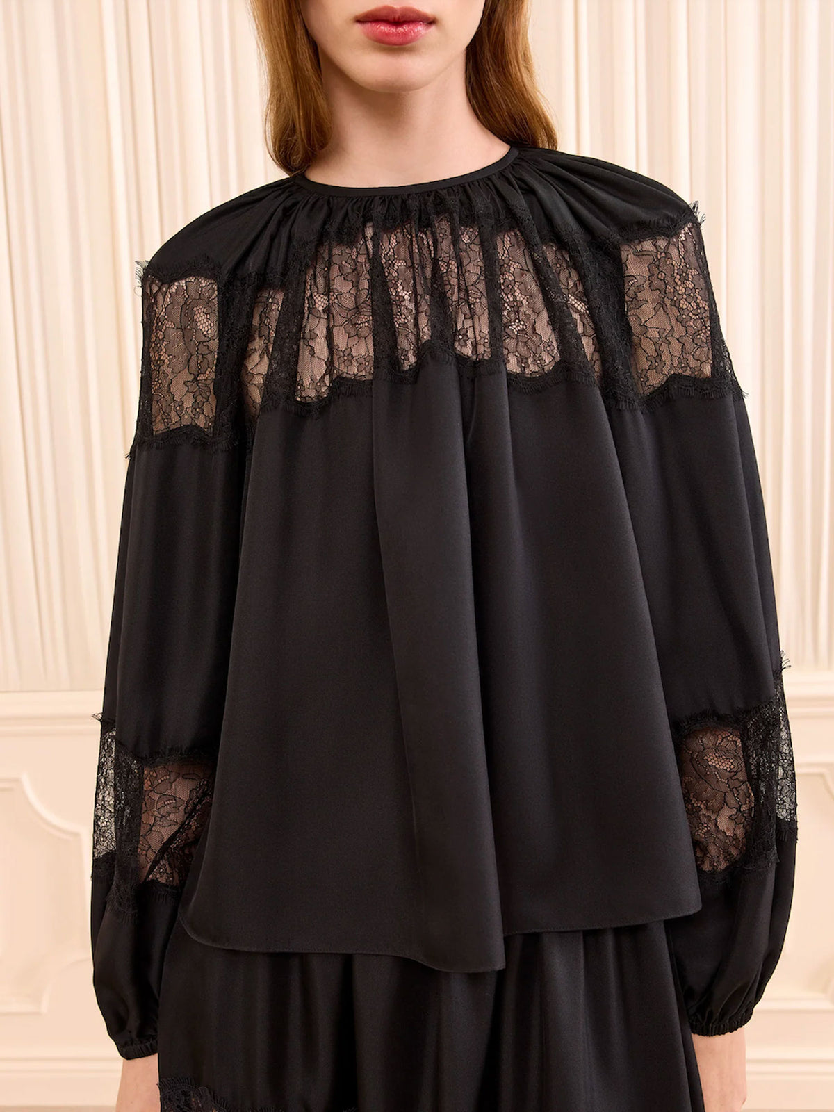 A model wearing a black long-sleeve blouse with sheer black lace panels across the chest and along the sleeves, set against a cream paneled wall.