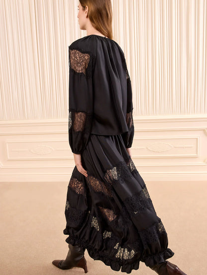 A woman wearing a black long-sleeve dress with sheer lace panels and gathered ruffles, shown from the back in an indoor setting with cream paneled walls.