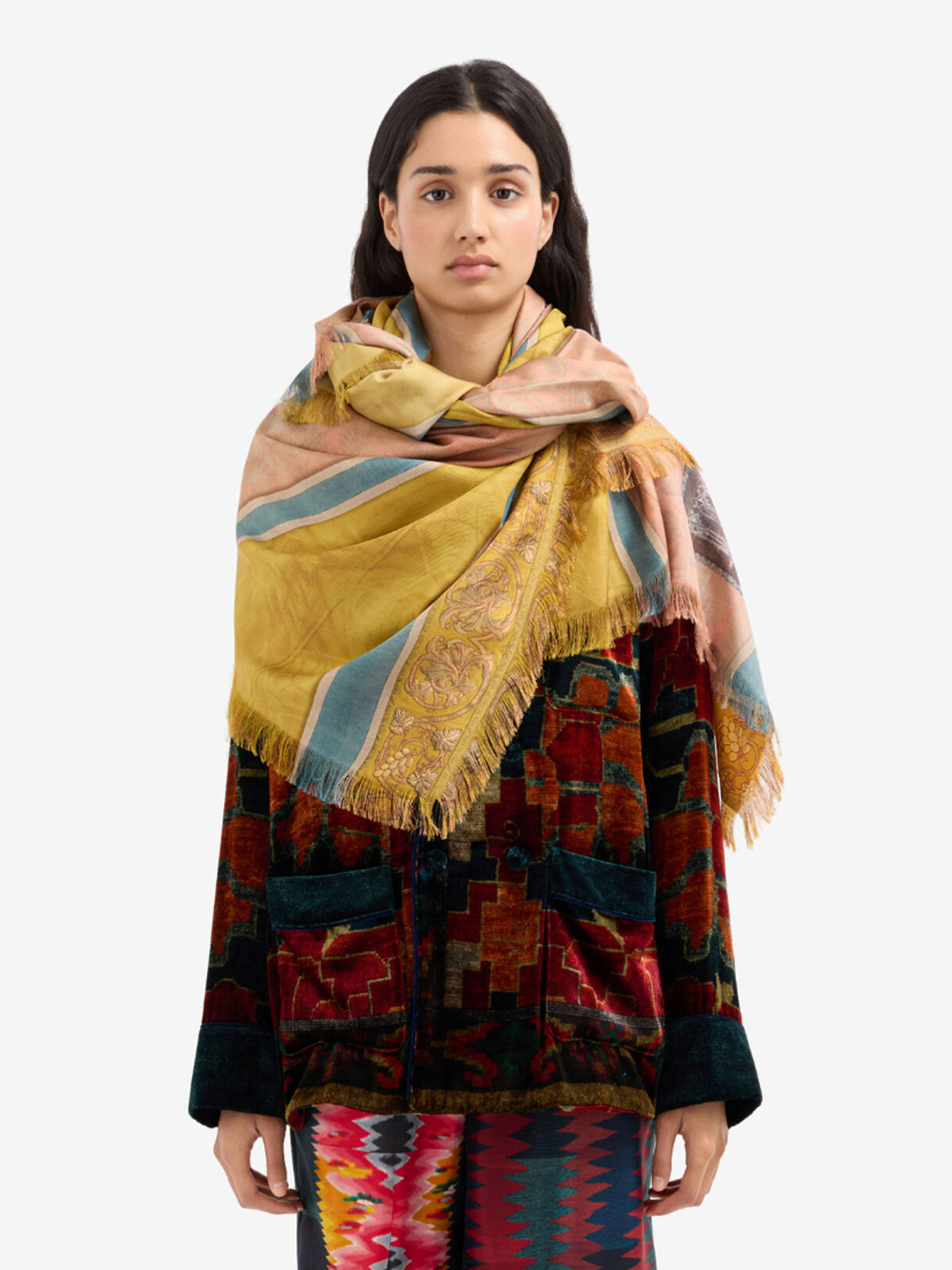 A model wearing a large, multicolored patterned scarf with fringe, draped over a dark patterned jacket against a plain light gray background.