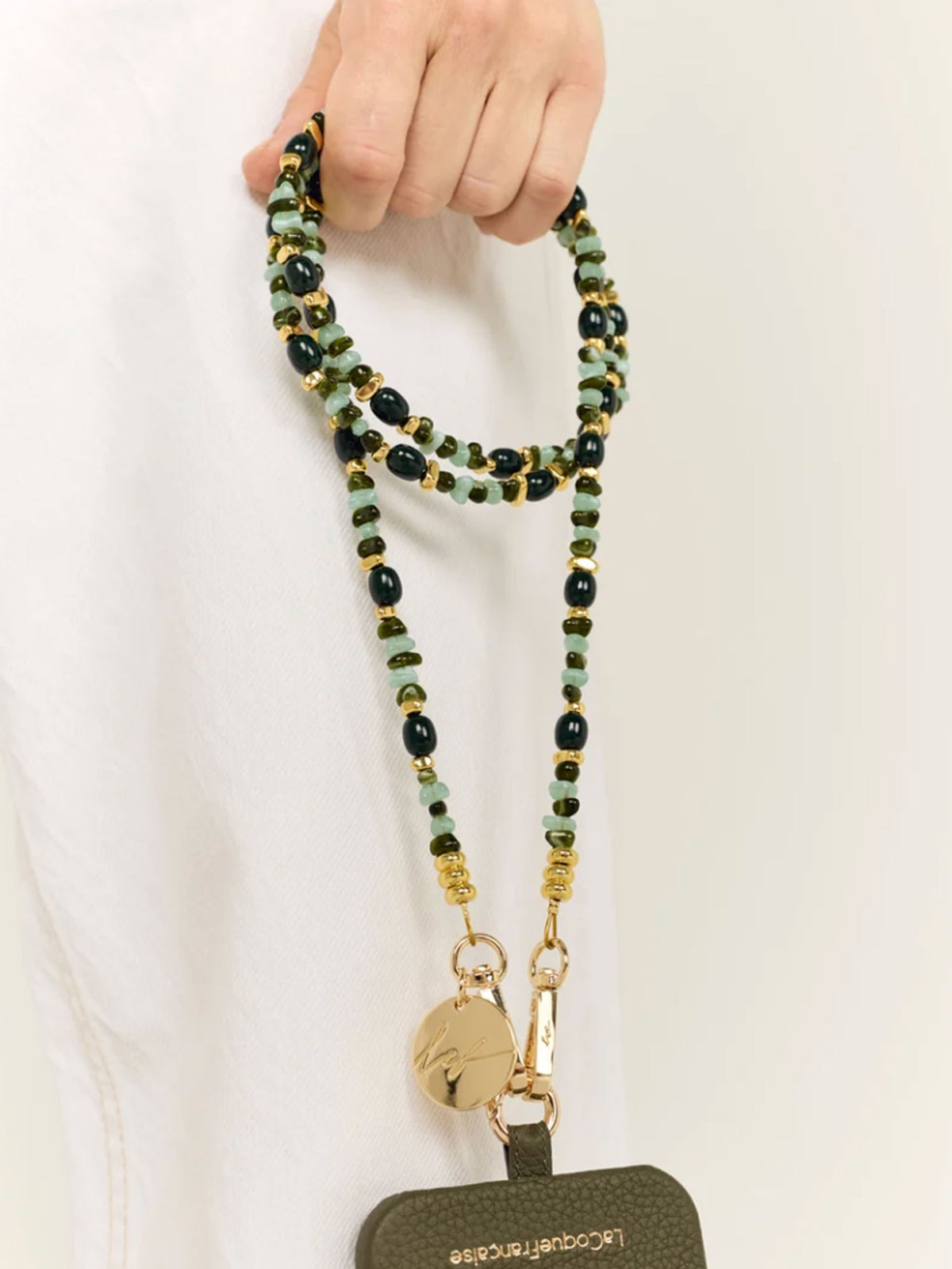 A close-up of a person holding a beaded lanyard strap with gold-tone hardware and a dark olive green pouch. The beads are primarily dark green, light mint green, and black, with small gold accents.