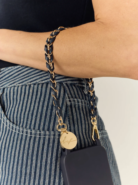 A close-up of a person's arm and waist wearing a black braided phone strap with gold-tone chain links, a round engraved gold charm, and a black phone case.