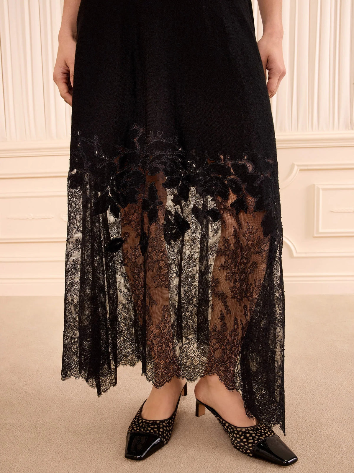 A black sheer lace midi skirt with floral applique and a flared hem, shown on a model wearing black studded pointed-toe heels against an elegant cream paneled wall.