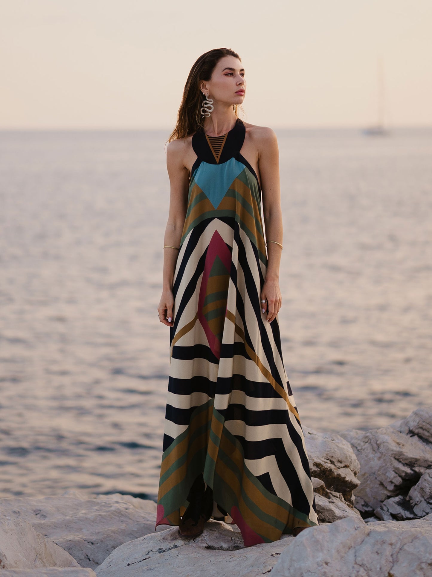 Woman in a colorful striped dress standing on rocks by the water.
